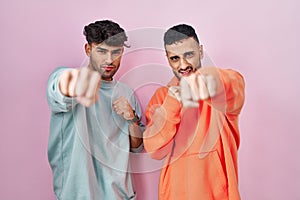 Young hispanic gay couple standing over pink background punching fist to fight, aggressive and angry attack, threat and violence