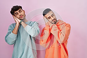 Young hispanic gay couple standing over pink background sleeping tired dreaming and posing with hands together while smiling with