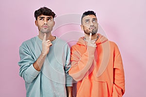 Young hispanic gay couple standing over pink background thinking concentrated about doubt with finger on chin and looking up