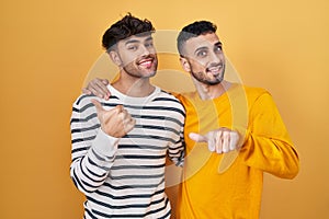 Young hispanic gay couple standing over yellow background pointing to the back behind with hand and thumbs up, smiling confident