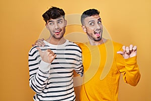 Young hispanic gay couple standing over yellow background smiling with happy face looking and pointing to the side with thumb up