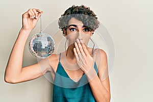 Young hispanic gay holding shiny disco ball covering mouth with hand, shocked and afraid for mistake
