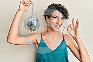 Young hispanic gay holding shiny disco ball doing ok sign with fingers, smiling friendly gesturing excellent symbol