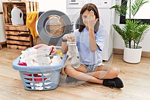 Young hispanic girl doing laundry holding socks covering one eye with hand, confident smile on face and surprise emotion