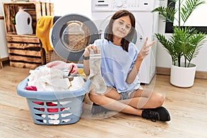 Young hispanic girl doing laundry holding socks smiling with happy face winking at the camera doing victory sign with fingers