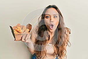 Young hispanic girl eating chicken wings scared and amazed with open mouth for surprise, disbelief face