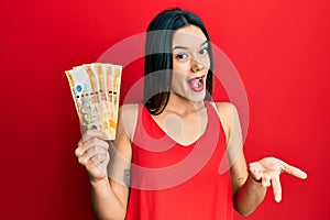 Young hispanic girl holding 500 philippine peso banknotes celebrating achievement with happy smile and winner expression with