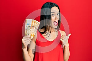 Young hispanic girl holding 500 philippine peso banknotes pointing thumb up to the side smiling happy with open mouth