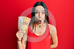 Young hispanic girl holding 500 philippine peso banknotes scared and amazed with open mouth for surprise, disbelief face