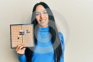 Young hispanic girl holding asian go game board looking positive and happy standing and smiling with a confident smile showing