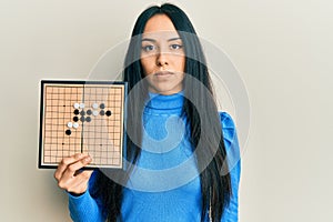 Young hispanic girl holding asian go game board thinking attitude and sober expression looking self confident
