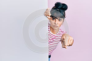 Young hispanic girl holding blank empty banner pointing with finger to the camera and to you, confident gesture looking serious