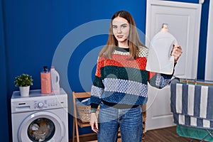 Young hispanic girl holding detergent bottle thinking attitude and sober expression looking self confident