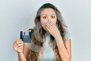 Young hispanic girl holding floppy disk covering mouth with hand, shocked and afraid for mistake