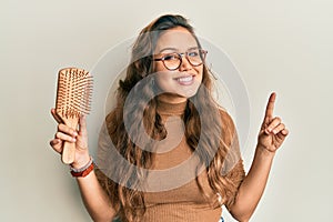 Young hispanic girl holding hair comb surprised with an idea or question pointing finger with happy face, number one