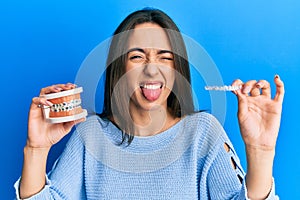 Young hispanic girl holding invisible aligner orthodontic and braces sticking tongue out happy with funny expression