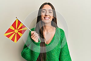 Young hispanic girl holding macedonian flag looking positive and happy standing and smiling with a confident smile showing teeth