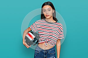 Young hispanic girl holding motorcycle helmet thinking attitude and sober expression looking self confident
