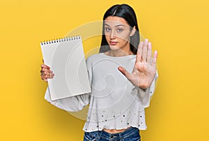 Young hispanic girl holding notebook with open hand doing stop sign with serious and confident expression, defense gesture