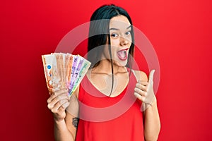 Young hispanic girl holding philippine peso banknotes pointing thumb up to the side smiling happy with open mouth