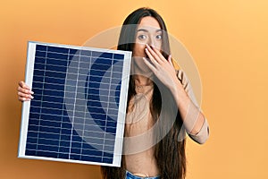 Young hispanic girl holding photovoltaic solar panel covering mouth with hand, shocked and afraid for mistake
