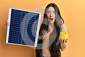 Young hispanic girl holding photovoltaic solar panel and piggy bank afraid and shocked with surprise and amazed expression, fear