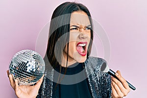 Young hispanic girl holding shiny disco ball and makeup brush angry and mad screaming frustrated and furious, shouting with anger