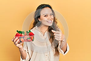 Young hispanic girl holding strawberries pointing fingers to camera with happy and funny face