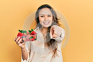 Young hispanic girl holding strawberries pointing to you and the camera with fingers, smiling positive and cheerful