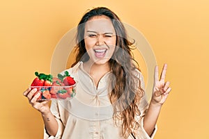 Young hispanic girl holding strawberries smiling with happy face winking at the camera doing victory sign with fingers