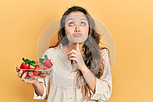Young hispanic girl holding strawberries thinking concentrated about doubt with finger on chin and looking up wondering