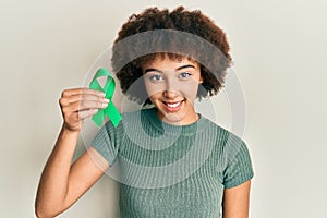 Young hispanic girl holding support green ribbon looking positive and happy standing and smiling with a confident smile showing