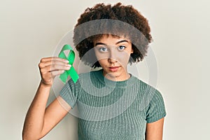 Young hispanic girl holding support green ribbon thinking attitude and sober expression looking self confident