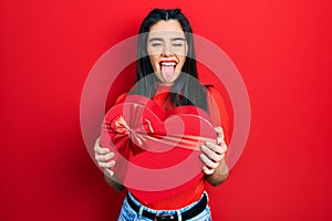 Young hispanic girl holding valentine gift sticking tongue out happy with funny expression