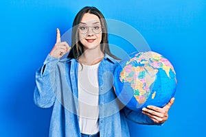 Young hispanic girl holding world ball surprised with an idea or question pointing finger with happy face, number one