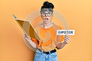 Young hispanic girl reading book and holding paper with hipster message in shock face, looking skeptical and sarcastic, surprised