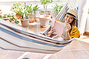 Young hispanic girl reading book lying on hammock at the terrace