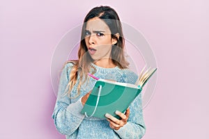 Young hispanic girl reading and writing book in shock face, looking skeptical and sarcastic, surprised with open mouth