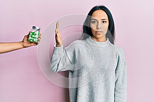 Young hispanic girl rejecting jar of pills with hand thinking attitude and sober expression looking self confident