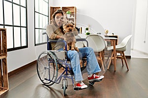 Young hispanic girl sitting on wheelchair at home doing ok gesture like binoculars sticking tongue out, eyes looking through