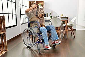 Young hispanic girl sitting on wheelchair at home peeking in shock covering face and eyes with hand, looking through fingers with