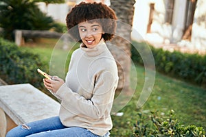 Young hispanic girl smiling happy using smartphone sitting on the bench at the park