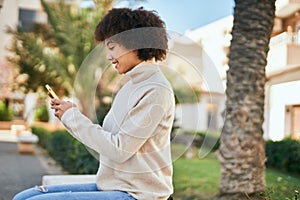 Young hispanic girl smiling happy using smartphone sitting on the bench at the park