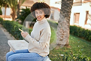 Young hispanic girl smiling happy using smartphone sitting on the bench at the park