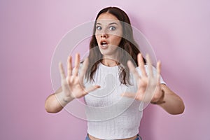 Young hispanic girl standing over pink background afraid and terrified with fear expression stop gesture with hands, shouting in