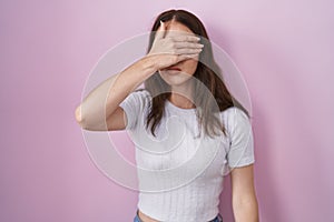 Young hispanic girl standing over pink background covering eyes with hand, looking serious and sad