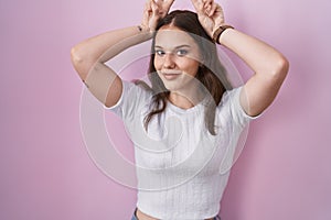 Young hispanic girl standing over pink background posing funny and crazy with fingers on head as bunny ears, smiling cheerful