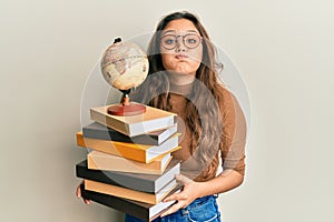 Young hispanic girl studying geography puffing cheeks with funny face