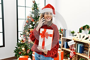 Young hispanic girl talking on the smartphone and holding gift standing by christmas tree at home