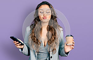 Young hispanic girl using smartphone and drinking a cup of coffee making fish face with mouth and squinting eyes, crazy and